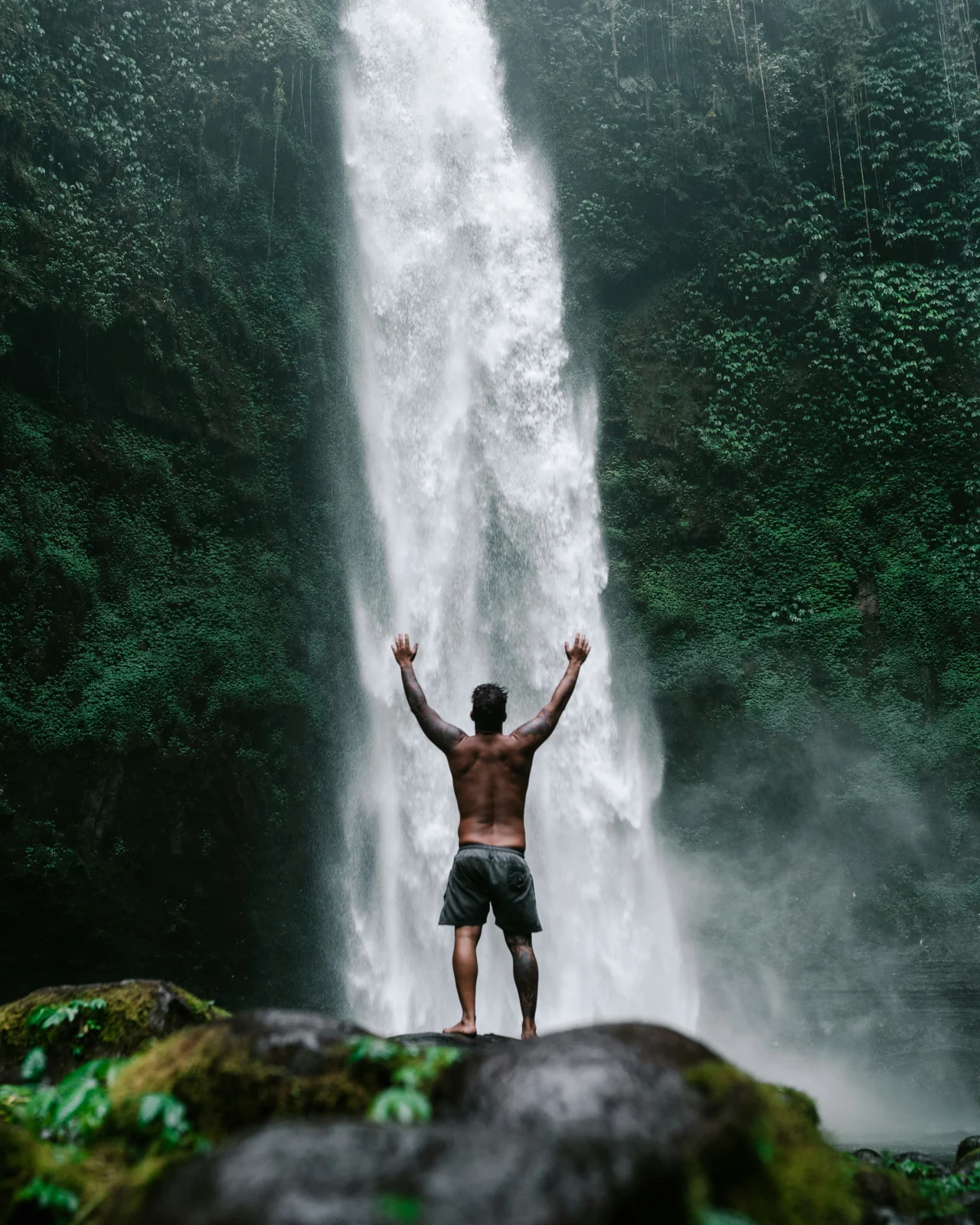 A traveller standing with arms raised at the foot of a jungle waterfall